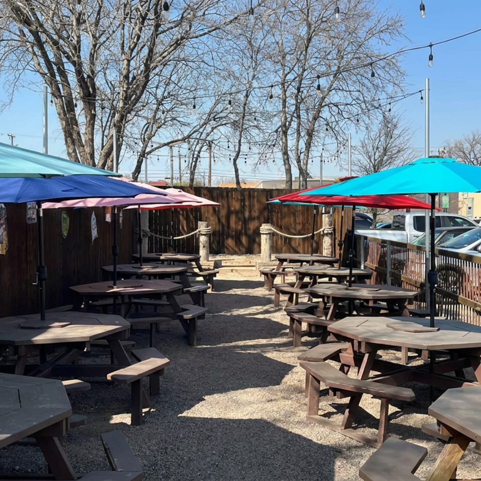 Outdoor restaurant patio with wooden picnic tables, colorful umbrellas, string lights, and parked cars beyond the railing.