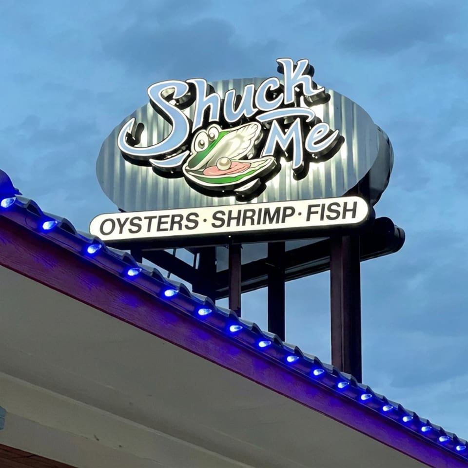Shuck Me seafood restaurant sign featuring oysters, shrimp, and fish above a blue-lit roof.