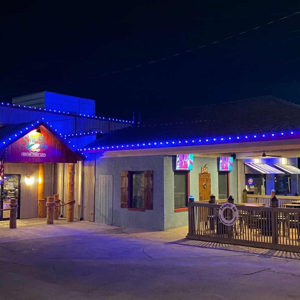 Nighttime exterior of a brightly illuminated restaurant with blue string lights, covered patio seating, picnic tables, and a glowing roadside sign.